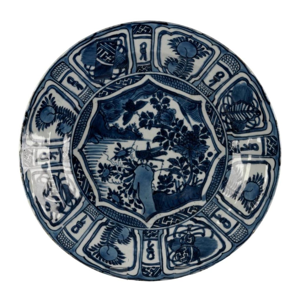 Round blue and white plate with dense, heavy blue designs divided into rectangles around the border. In the middle is a cricket on a branch by a pond.