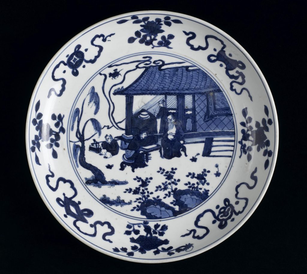 Round blue and white plate with a border of squiggly designs and a central scene of four characters seated in a garden outside a house.