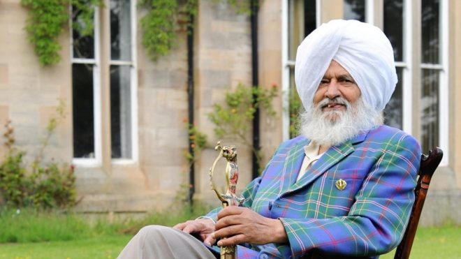 Man seated in a wooden chair on a grassy lawn, wearing a white turban and tartan jacket and holding an ornate sword.