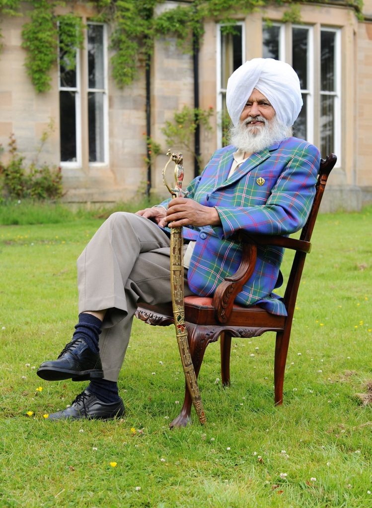 Man seated in a wooden chair on a grassy lawn, wearing a white turban and tartan jacket and holding an ornate sword. 