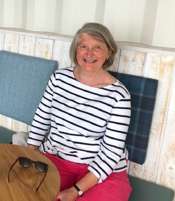 Woman in a white and black striped shirt and pink trousers sits at a small wooden table with a back bench.