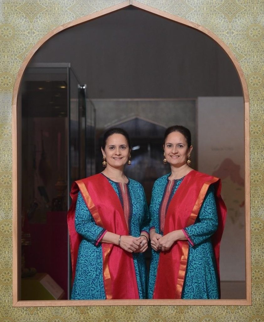 Two women in red sashes and blue dresses with floral patterns stand under an arch within a gallery space.