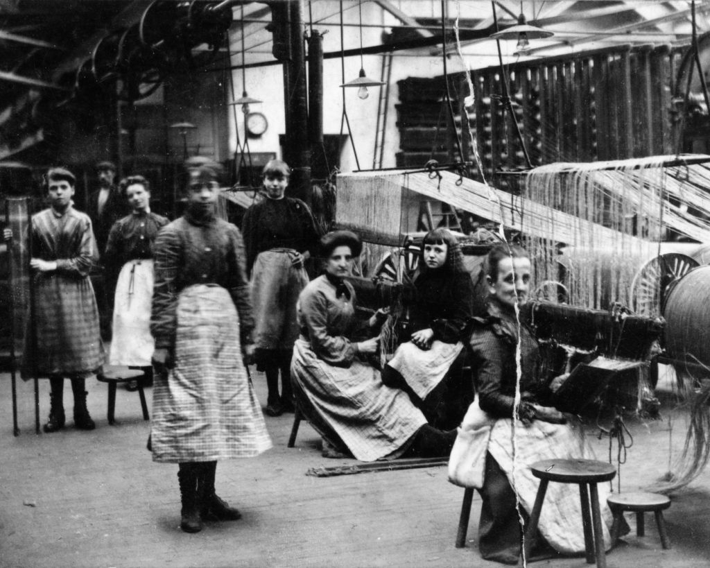 Black and white photo of a jute mill with women standing and sitting around the mills.