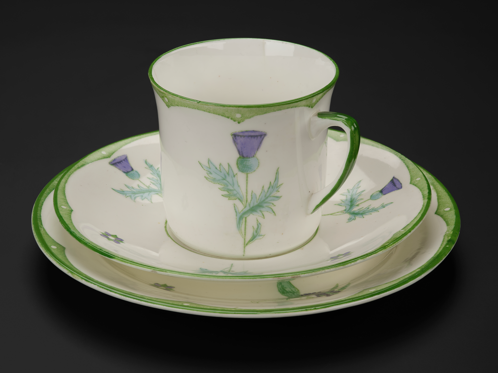 A cup on a saucer on a plate. They're white with pale green edging and thistles. And a thistle is on the cup.