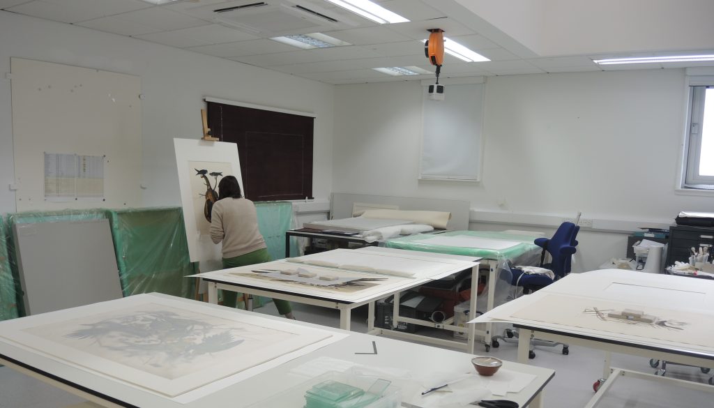 Colour photo of the interior of a modern conservation studio with white tables in the foreground and a woman working at an easel in the background.