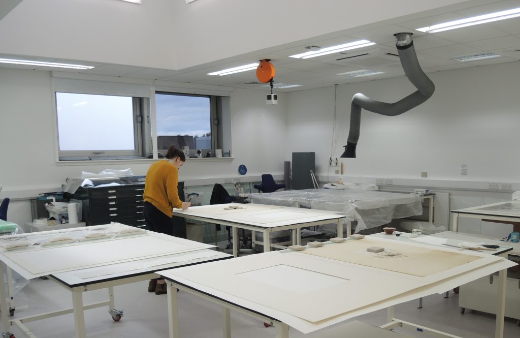 Colour photo of the interior of a modern conservation studio with white tables in the foreground and a woman working at one of the tables in the background.