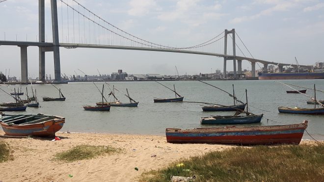 View from a sandy shore out to a wide river busy with small fishing boats and spanned by a large bridge.