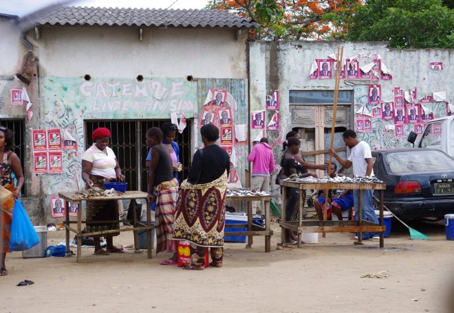 Market stalls set up in front of stone walls covered with posters. Merchants sell fish to eager customers wearing capulana.