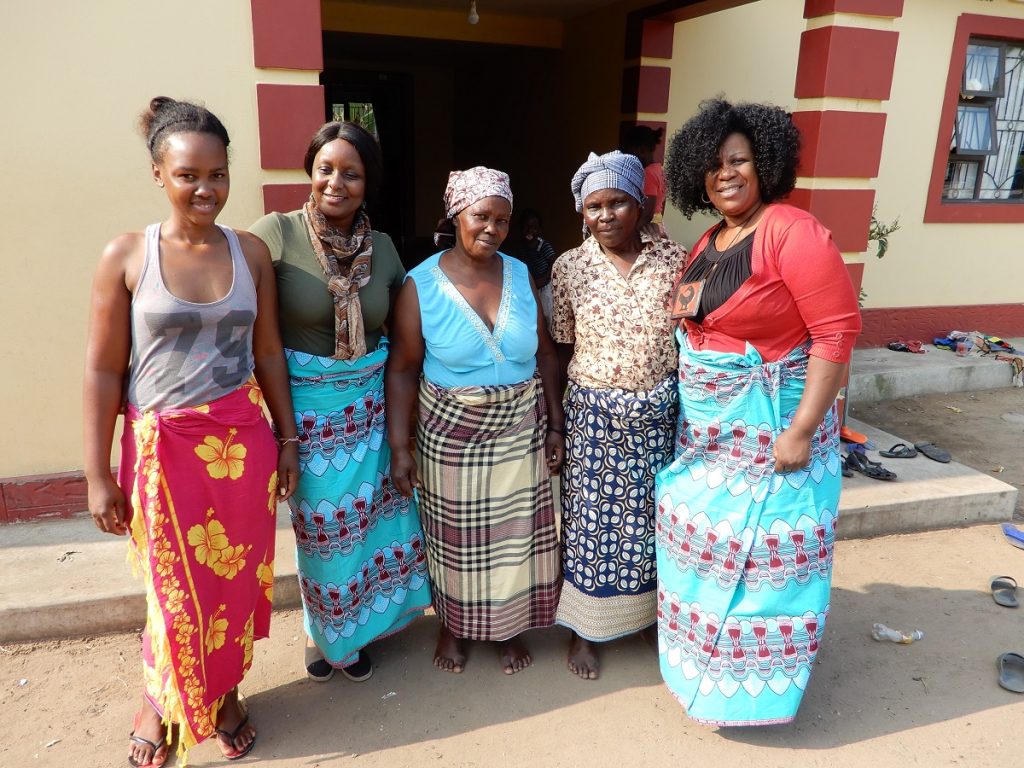 Intergenerational group of five women dressed in vibrant, colourful capulana stand together outside a red and yellow building.