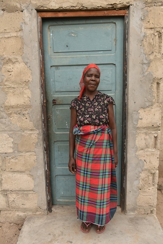 Woman dressed in red and green capulana resembling tartan stands outside in front of a reen door.