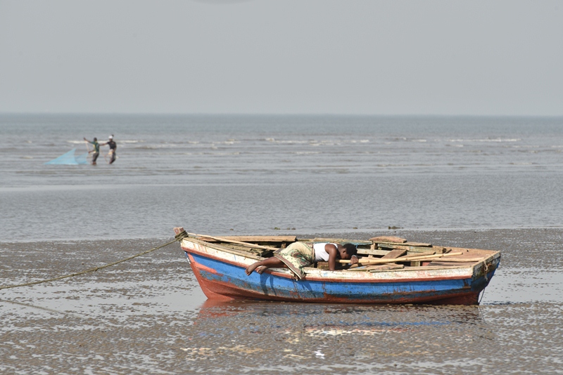Small boat with wooden planks across it and a woman lying on top of it alongside a sea stretching to the horizon.