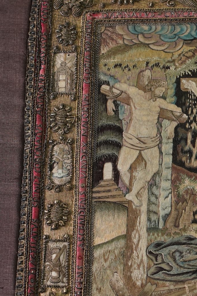 Detail of the border of the textile, with gold embroidery and sequins on a red satin ground, and an abbreviated Latin prayer describing Christ’s Passion and death. The small images show the Instruments of the Passion.