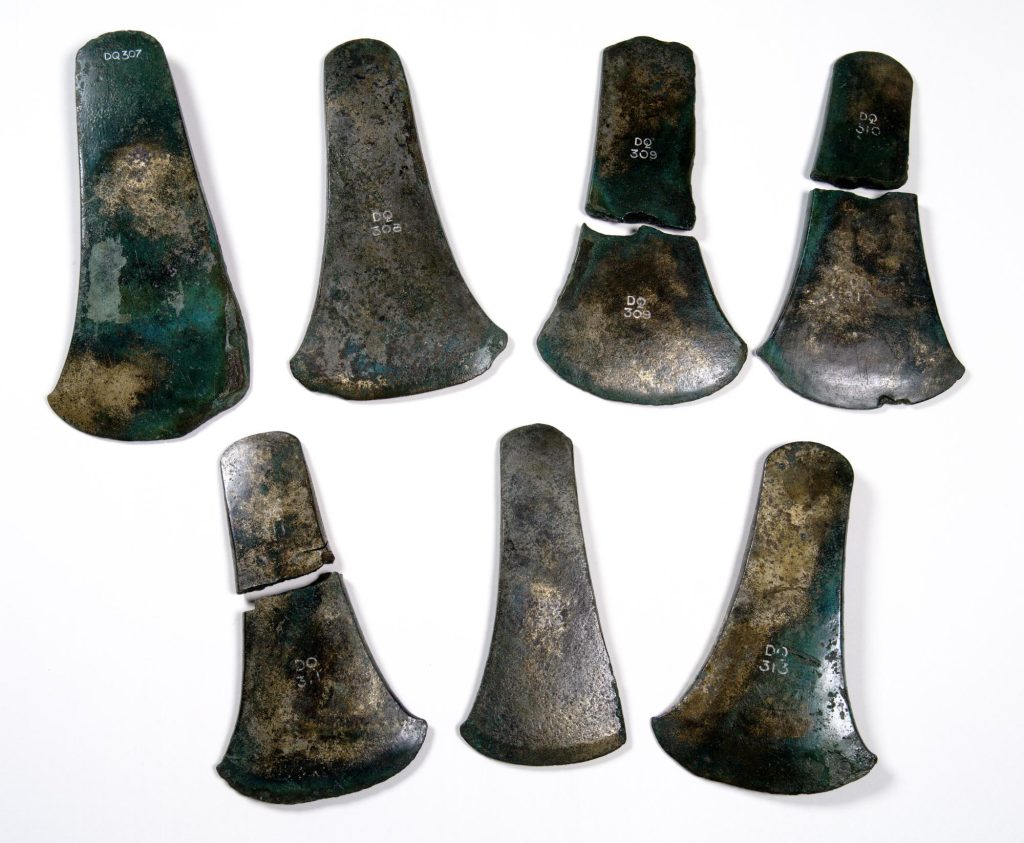 Set of seven bronze axes, coloured dark grey with yellow-green discolouration. Three are snapped in half.