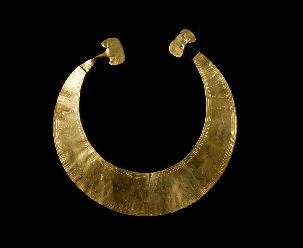 Flat, crescent-shaped gold object resembling a necklace, open at the top. Black background.
