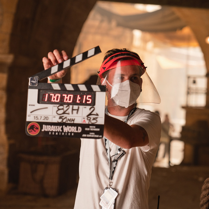 A man in a face covering stands with a clapperboard ready to snap.