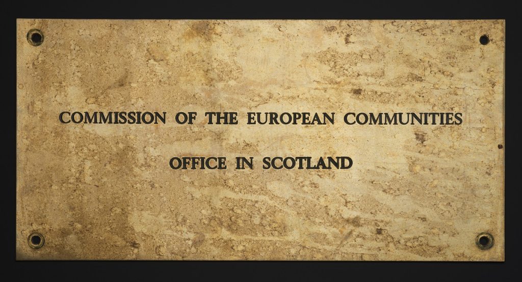 Weathered brass sign, perfectly rectangular, with small, black font across its centre reading in all caps, 'Commission of the European Communities Office in Scotland'.