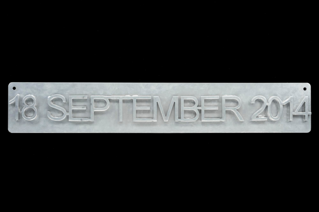 Grey steel sign rectangular and narrow with slightly rounded corners viewed straight on, with raised text across it reading '18 SEPTEMBER 2014'.
