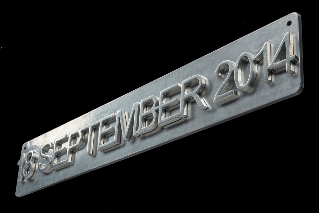 Grey steel sign viewed at a 45 degree low angle, rectangular and narrow with slightly rounded corners. Raised text across it reading '18 SEPTEMBER 2014'.
