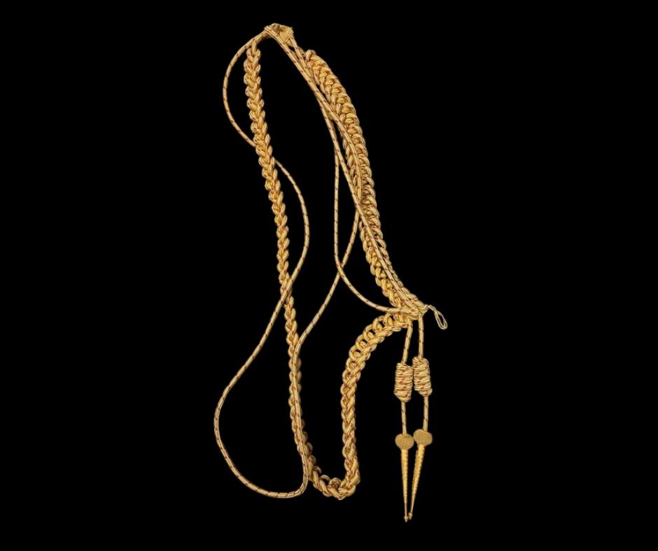 Golden rope-like golden cord forming a tall oval with tassels hanging down on the right side. Resting on a grey surface.