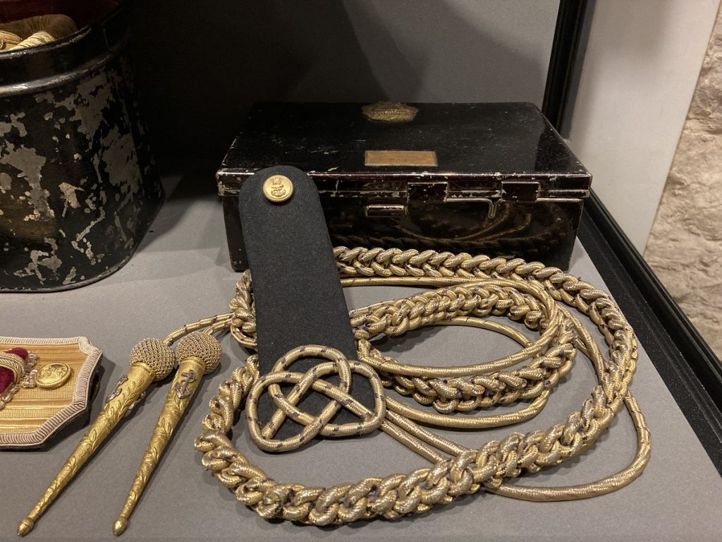 Golden rope-like cord with long, triangular points alongside a black case within a glass museum display case.
