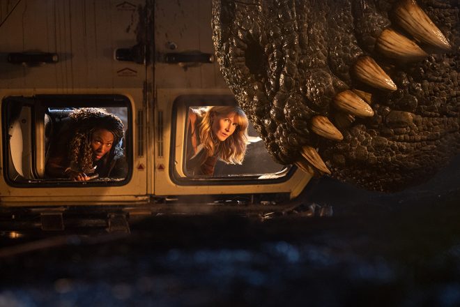 Still from Jurassic World Dominion showing two women in an upturned van staring our at the snout of a large dinosaur with sharp teeth.