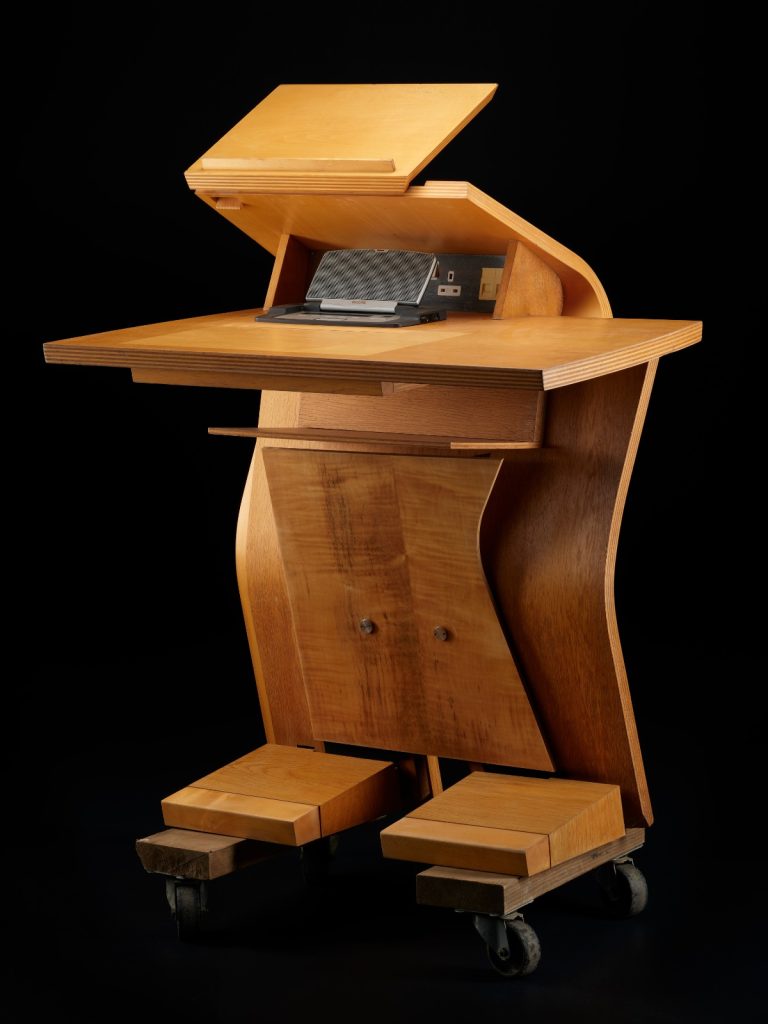 Light brown wooden desk shaped like a humanoid robot, with two blocky 'feet', a tall, curved centre, and two flat surfaces at the top.