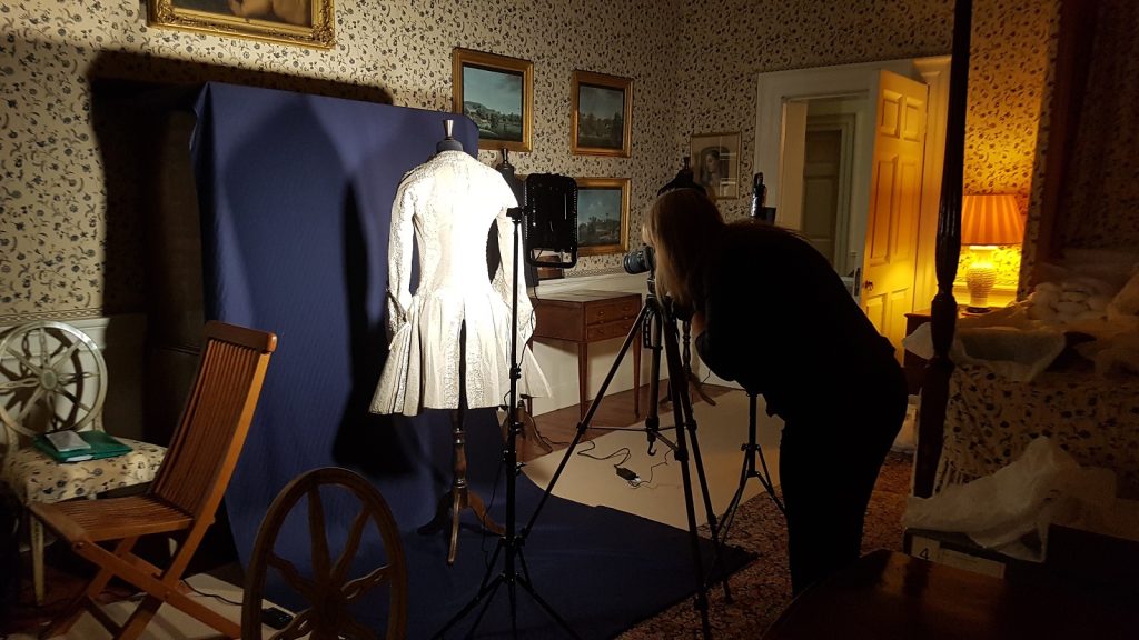 A woman photographs a brightly lit white coat against a blue screen inside a sumptuous 18th century room.