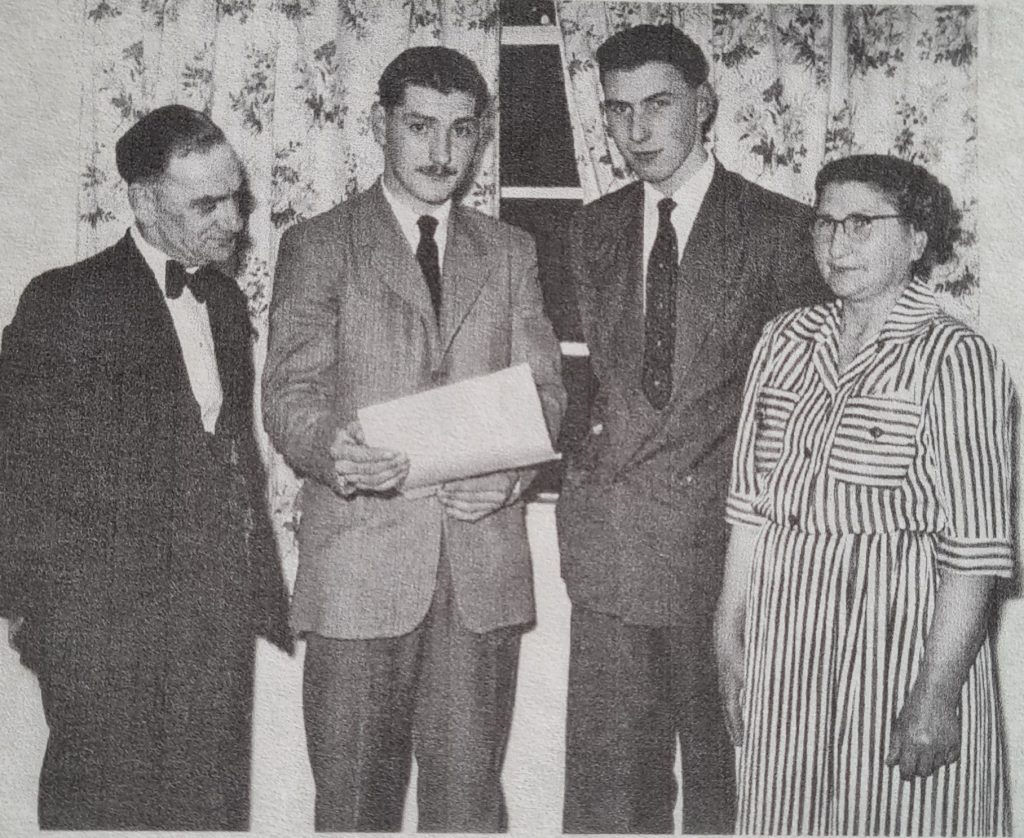 Black and white picture of four people, a middle-aged man on left, two young men in centre, and middle-aged woman on right, in 1950s suits. The young man in centre holds a piece of paper. They look proud. 