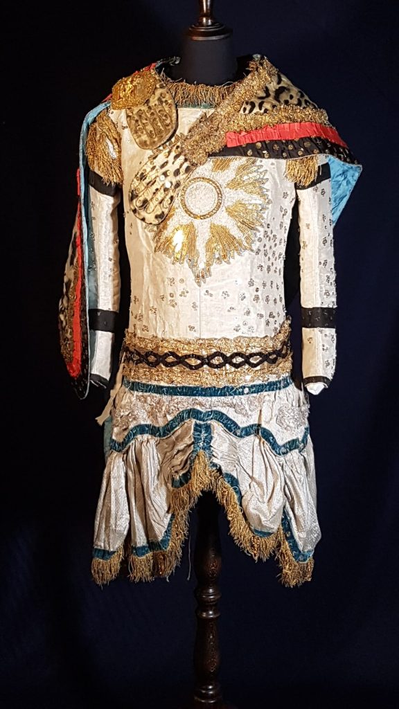 Decadent costume of a 'Carthaginian knight'. Cream coloured, with golden sun on the chest, leopard skin cape across the shoulder, blue-trimmed skirt, and glittering patches across the torso.