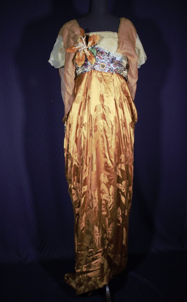 Evening dress on a stand against a blue background. Pink stripes form at the shoulders, a large orange flower is stuck on the front, and a shimmering golden long skirt runs from above the waist to the floor.