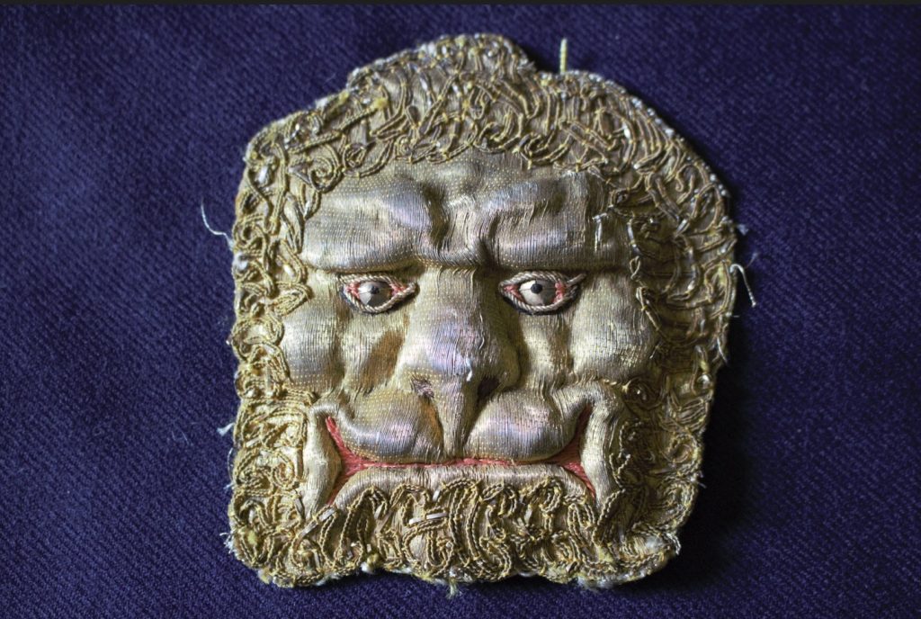 Embroidered depiction of a lion's head, looking more like the classic 'Green Man' than a lion, with fleshy features, uncanny eyes, and gold-embroidered mane resembling curly hair with mutton chop facial hair.