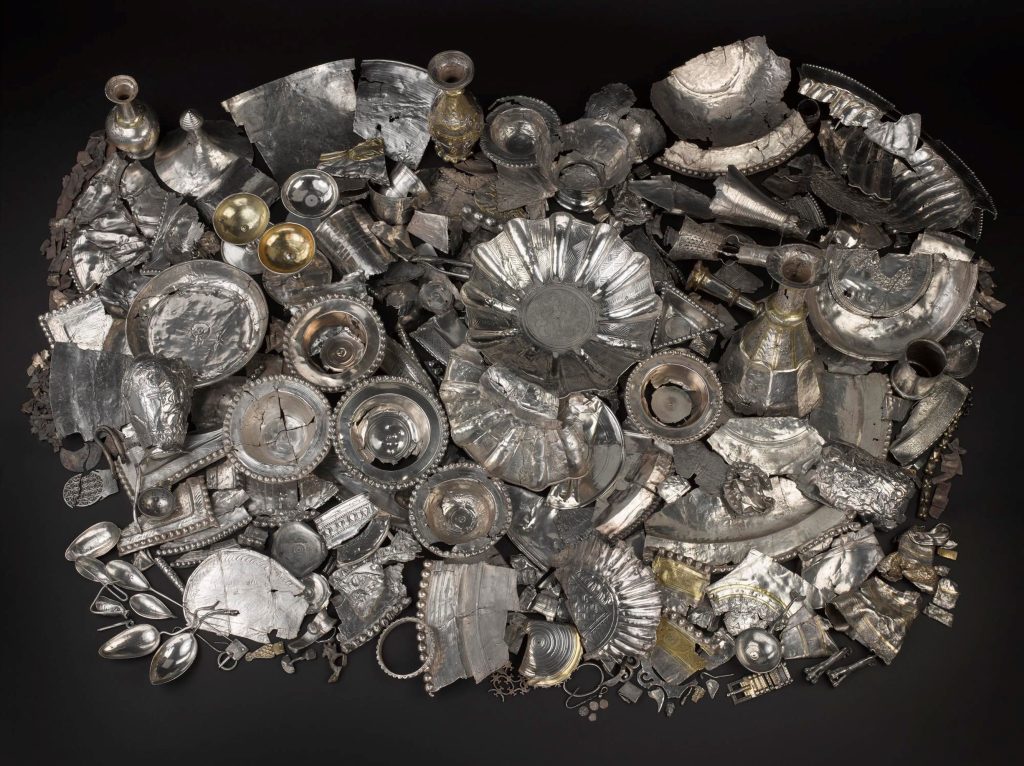 Massive pile of hundreds of silver objects viewed from above on a black surface. Objects include large plates and bowls, drinking vessels, spoons, small sculptures, and many shards.