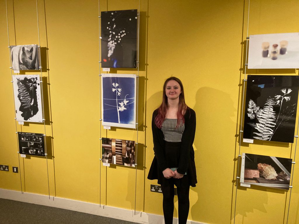 Colour photo of a girl standing in front of a yellow wall with photos display on it.