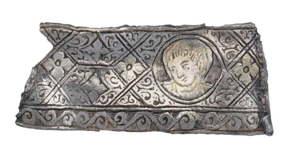 Rectangular fragment densely decorated with wave and diamond patterns. Within a circle is a childlike face looking bemused.