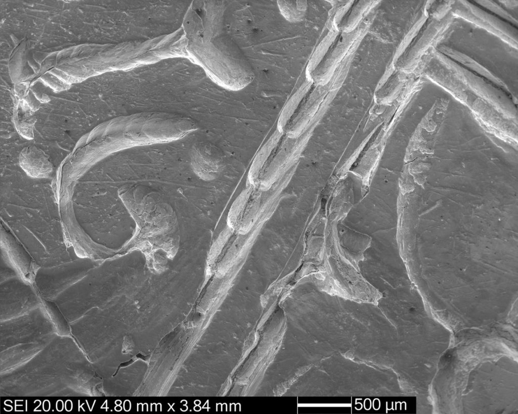 Electron microscope view, in bright silvery-grey, showing miniscule marks resembling ropes or hooks on a silver surface.