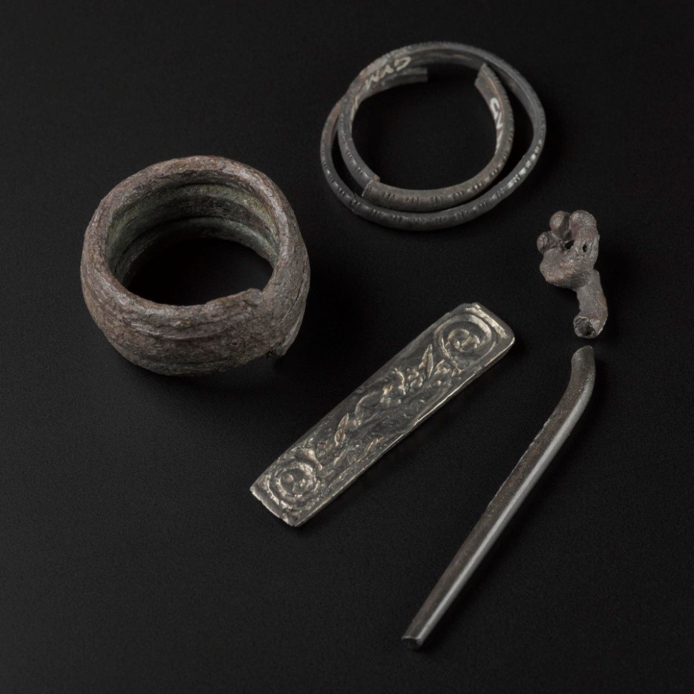 Four silver objects on a dark surface. At top, two rusted coils with faint engravings. In middle, a rectangular sheet with leaf design. On right, a long, thick pin.