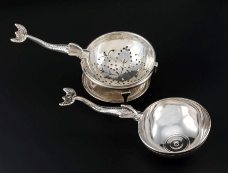 Two silver spoons side-by-side with elongated handles resembling thin dolphins. The one at top-left has been turned into a tea strainer. 