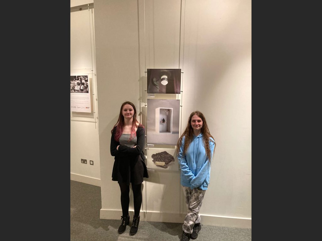Colour photo of two girls standing in front of a display of photos on a wall.