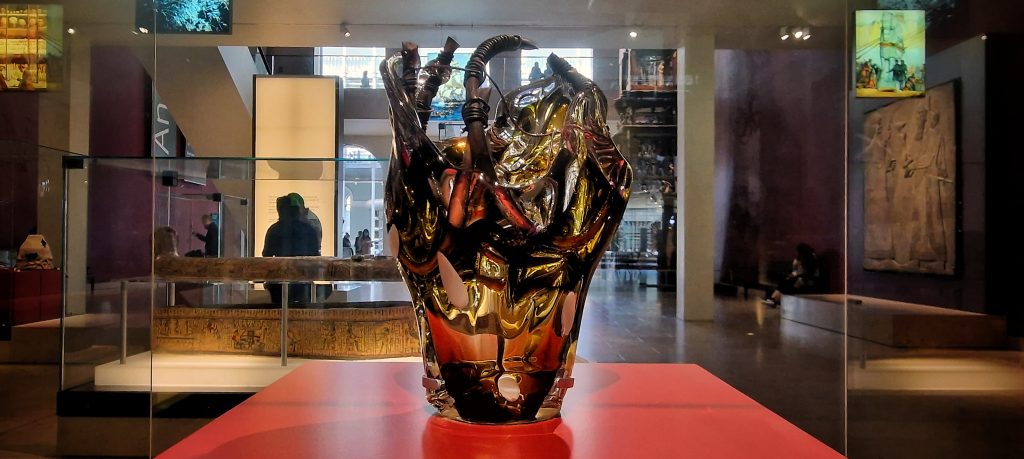 A glass and metal sculpture in a glass case on a red plinth.