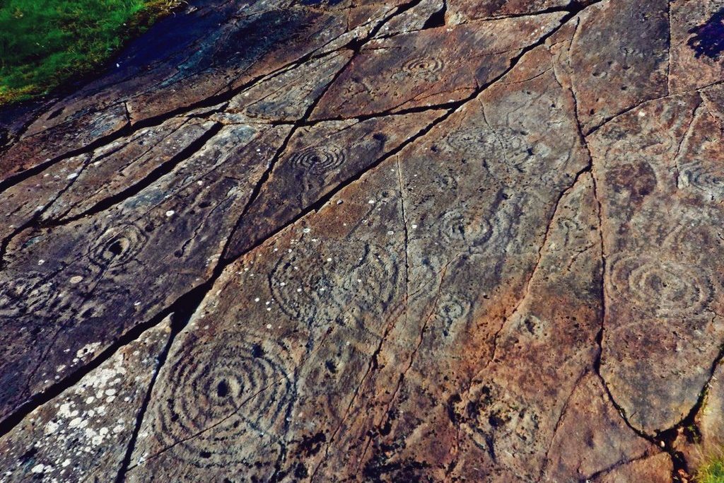 Sloping rocky outcrop, grey-orange in colour, decorated with large cup-and-ring rock art. Some of the cups have seven or more rings surrounding them in concentric circles.
