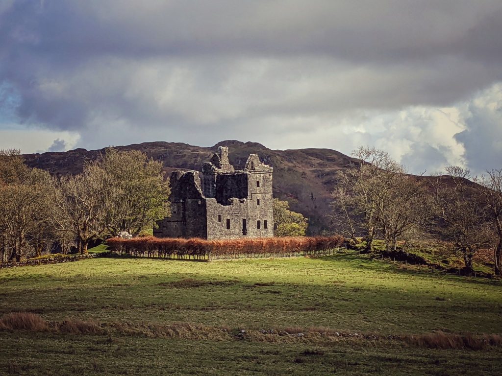 A ruinous grey castle, its tower roofless, stands within an enclosure of red shrubs set against rocky hills and flanked by autumnal trees. Light falls on the castle, leaving the surrounds in shadow.