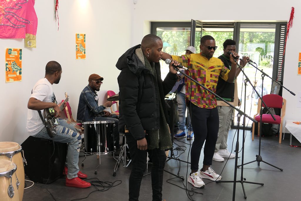 A group of Black, male musicians playing instruments and singing in a workshop space.