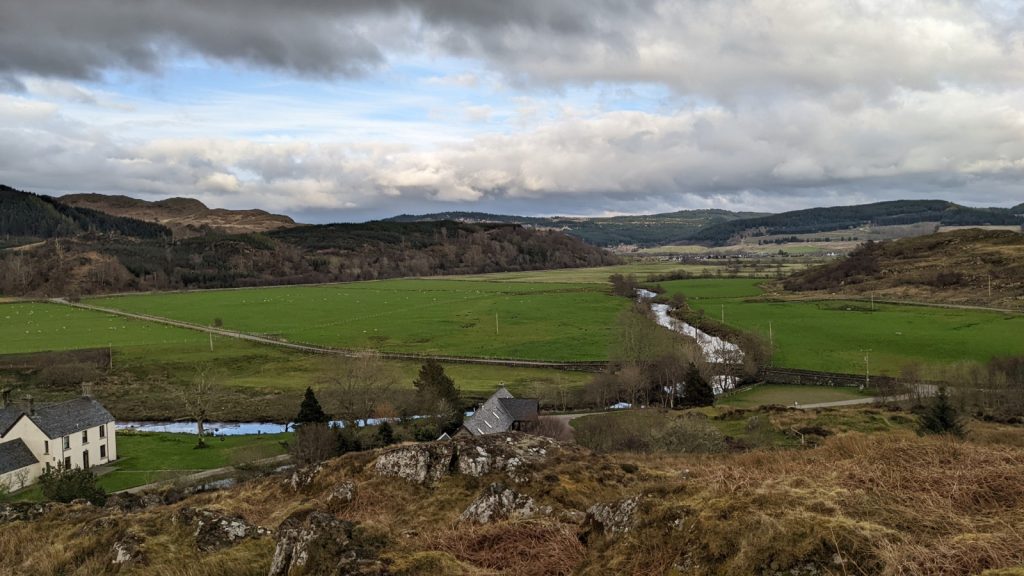 Sprawling view from atop Dunadd over a modest river winding its way through green fields and underneath an arched stone bridge. Two cottages are in the foreground, with forest hills continuing in the background.