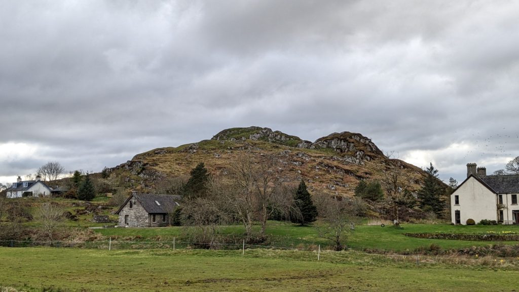A rocky crag with two distinct humps rises up from a grassy area. Three small cottages surround it. Heavy grey skies loom overhead.
