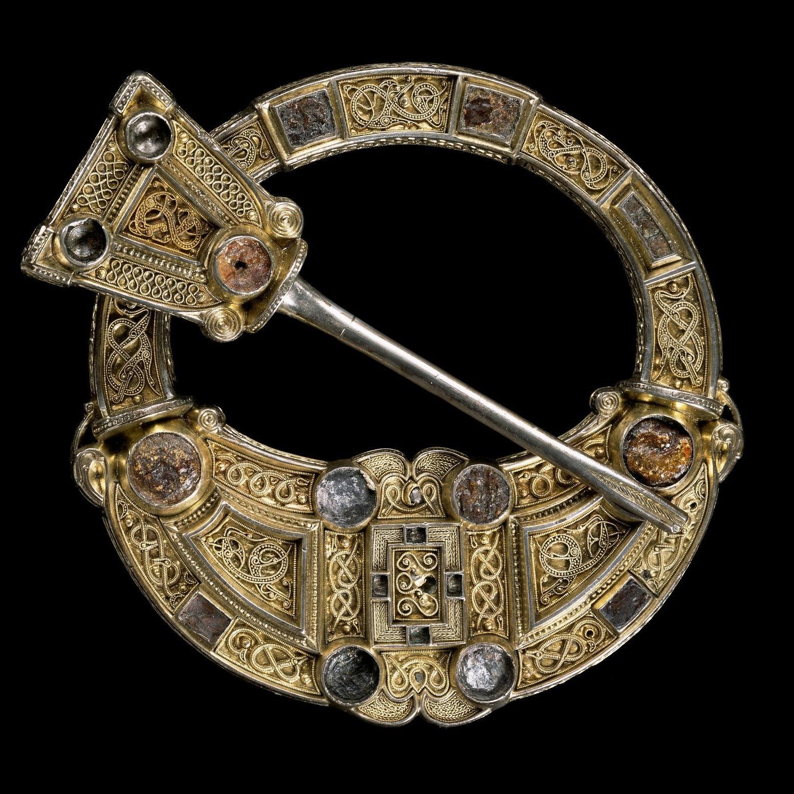 Stunningly ornate gold brooch, circular with a hollow top half and quill-like pin cutting diagonally across. Dense knot patterns and gemstone fittings cover its whole surface.