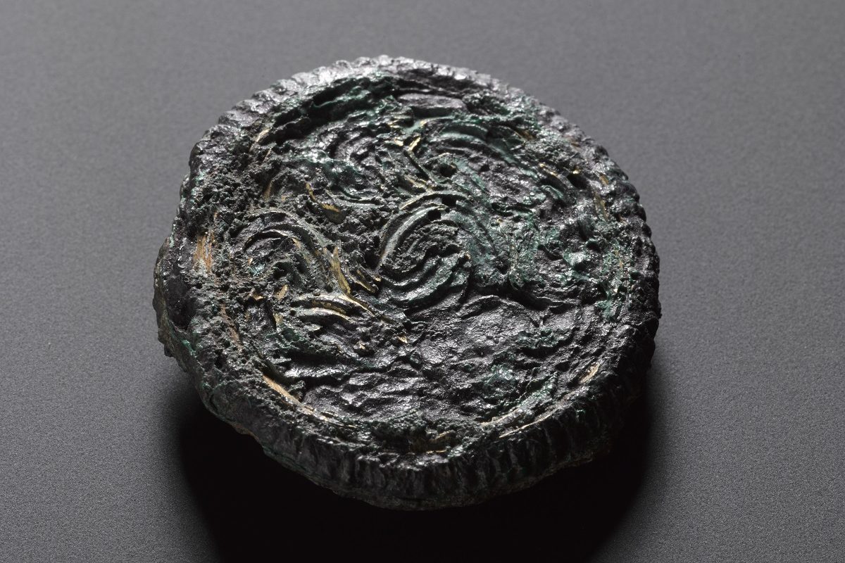 A now misshapen and blackened disc, decorated with swirling patterns that resemble ropes covered in oil. 