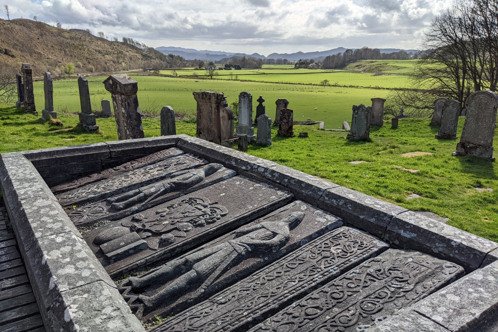 A horizontal stone enclosure on the ground of a graveyard containing seven grave slabs decorated with swords, vine patterns, and armoured warriors. Above is a serene, sunlit glen.