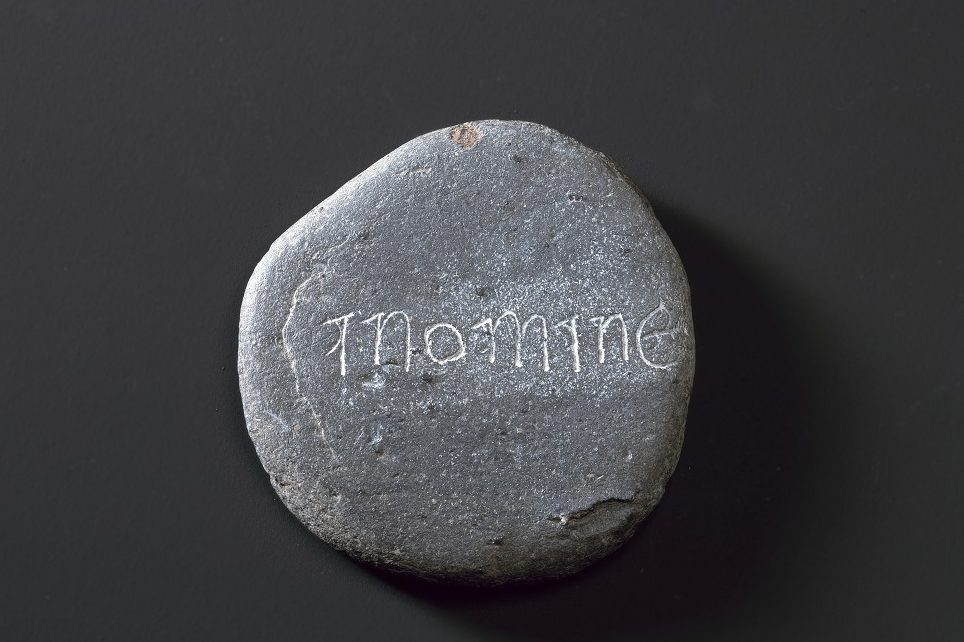 A roughly circular grey stone carved with white letters reading 'I N O M I N E', meaning 'in the name' in Latin.
