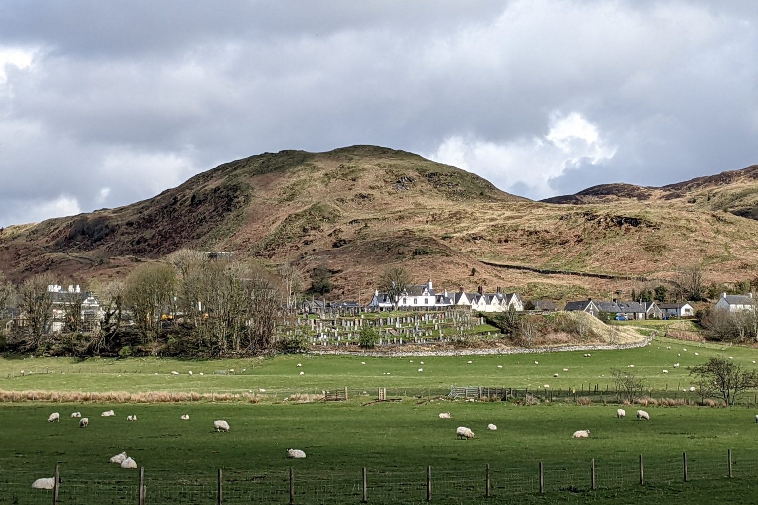 A small village punctuated by a stone church and white cottages stands on a natural earthen mound. Sheep graze green fields in the foreground, with bare hills rising immediately behind and above the village.
