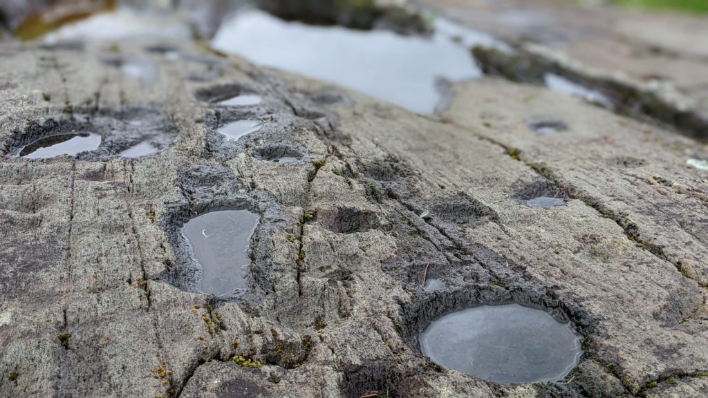 Closeup horizontal view of a stone surface adorned with various kinds of rock art, each filled with rainwater. Various cup marks along with an axehead shape.